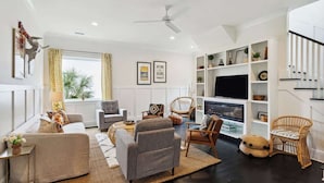A modern living room with a sofa, armchairs, a coffee table, and a TV. There is a bookshelf with decorative items and a staircase to the right. - Folly Oasis | Beachside Home w. Pool + Parking (Folly Beach)