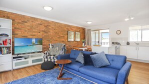 A living room with a blue sofa, a television, a brick wall, and a kitchen area. - Moola Palms - Pet Friendly (Hawks Nest)
