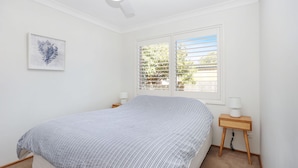 A bedroom with a bed, a window with blinds, a bedside table with a lamp, and a wall-mounted artwork. - Moola Palms - Pet Friendly (Hawks Nest)