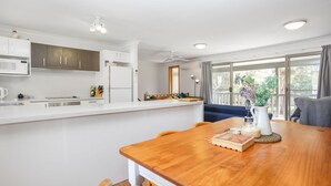 A modern kitchen with white cabinets, a microwave, and a refrigerator. There is a dining area with a wooden table and chairs, and a window with a view of greenery outside. - Moola Palms - Pet Friendly (Hawks Nest)