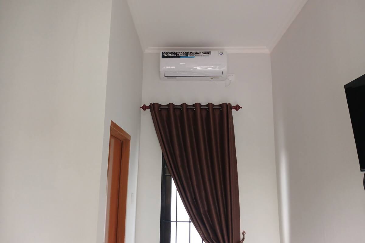 Une pièce avec un climatiseur fixé au plafond, une fenêtre avec un rideau marron et une porte en bois.