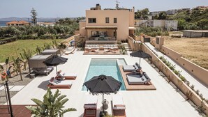 Un área de piscina con sillones de descanso y sombrillas, una piscina y un edificio de techo plano al fondo.