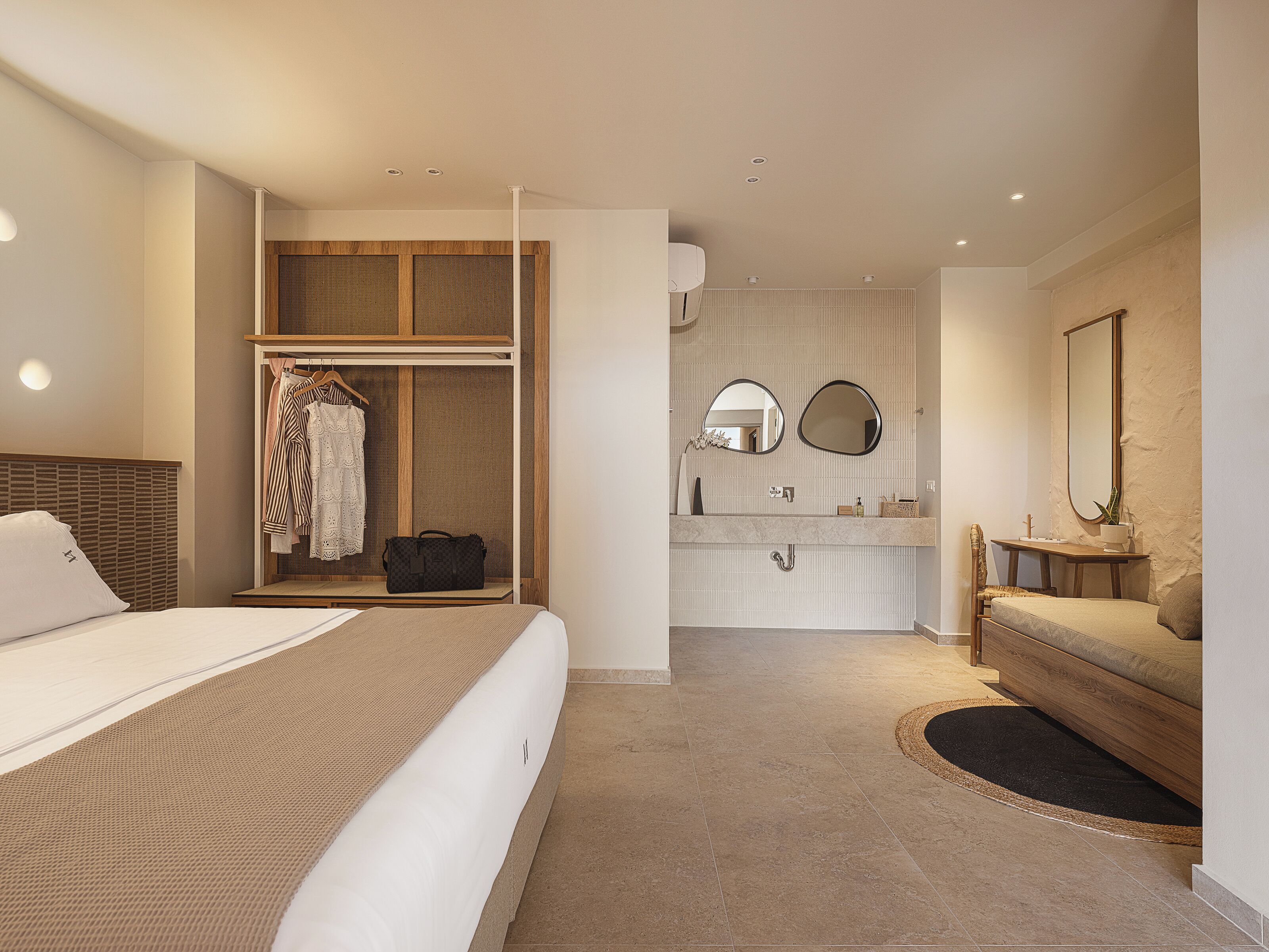 Une chambre d’hôtel moderne dotée d’un grand lit, d’un dressing, d’une salle de bain avec un meuble-lavabo et un miroir, et d’un petit coin salon.