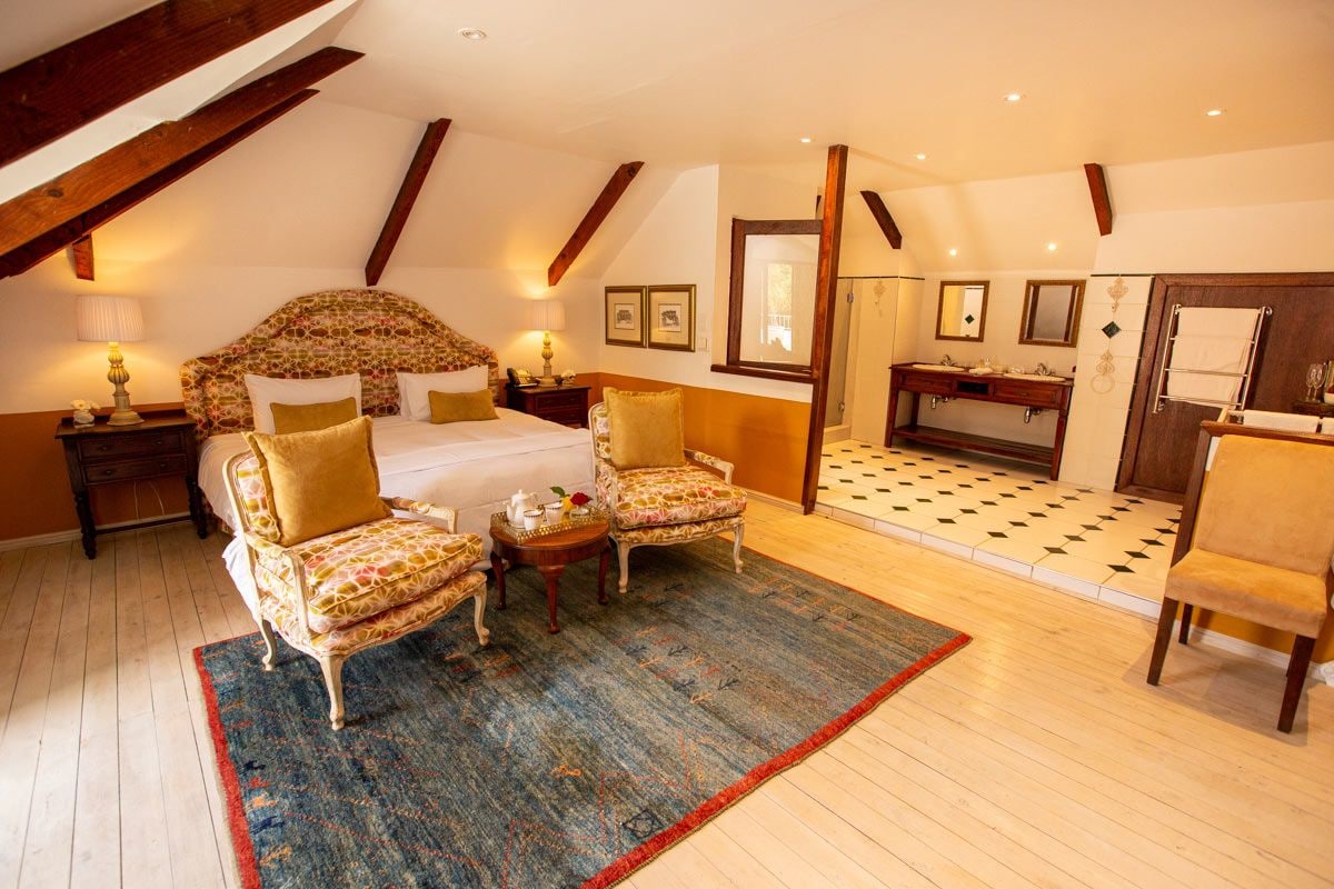 Une chambre à coucher avec un grand lit, deux fauteuils, une petite table avec une lampe, une commode en bois et une salle de bain avec un lavabo et un miroir.
