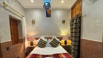 Habitación de hotel ordenada con una cama, lámparas de noche, una lámpara decorativa en el techo y una mesita de madera con un control remoto.