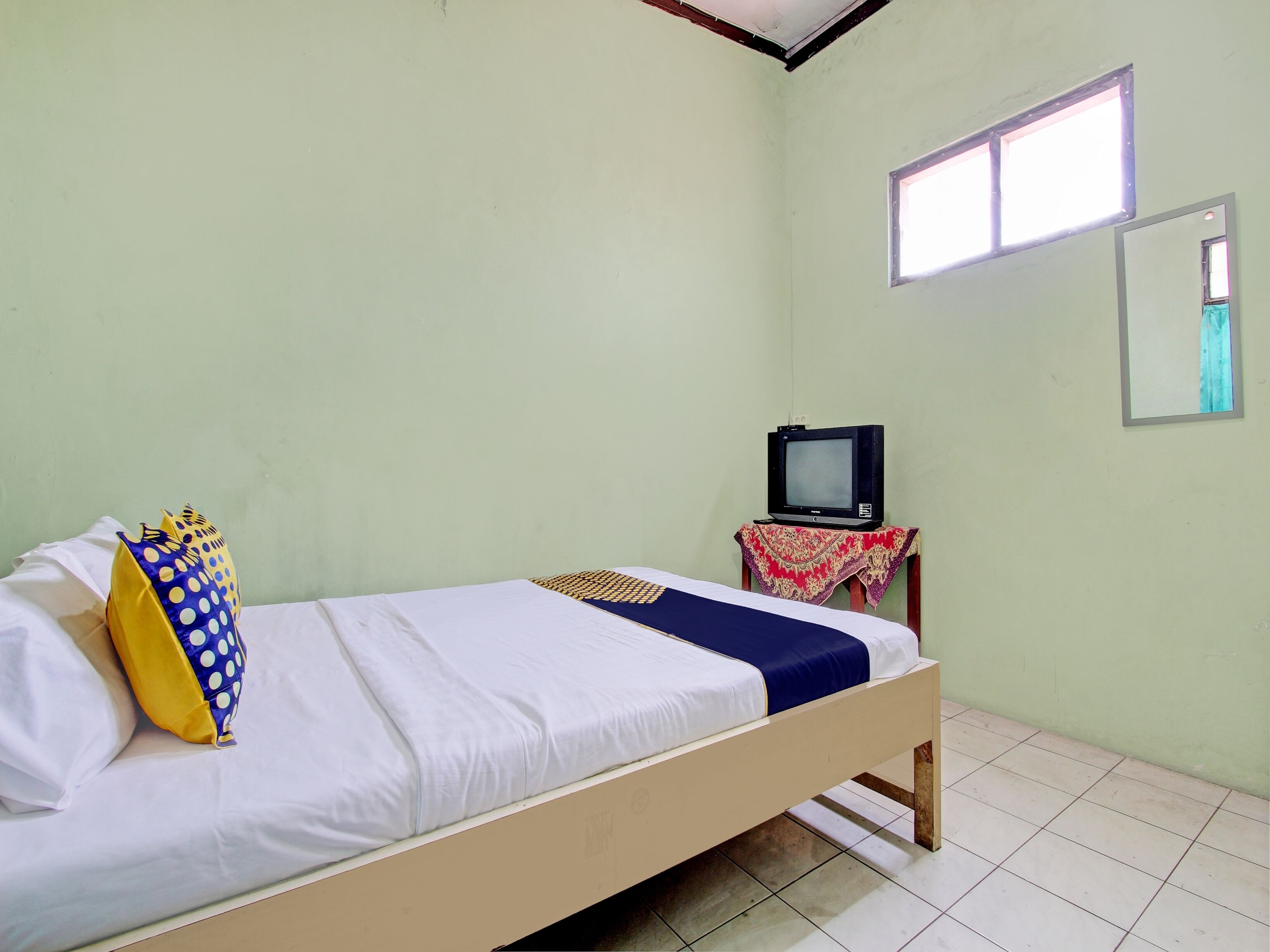 Una cama individual con ropa blanca y una almohada azul y amarilla, una mesita con un televisor, y una ventana que da a un cielo azul.