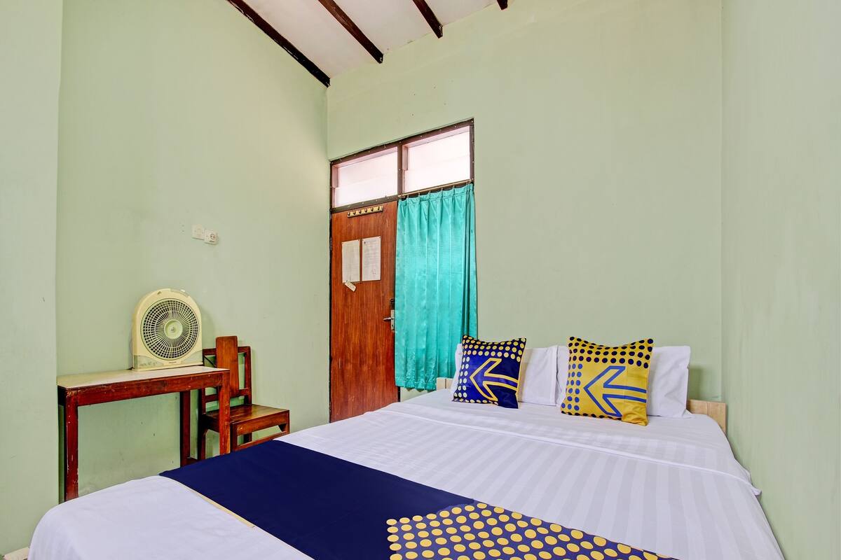 Una habitación con cama, escritorio, silla, ventana con cortinas y ventilador de pared.