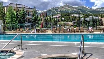 Un área de piscina con sillones de descanso y un jacuzzi, rodeado de edificios y montañas.