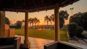 Een overdekt buitengebied met een geruit vloertje, lounge stoelen en uitzicht op palmbomen en een zonsondergang.