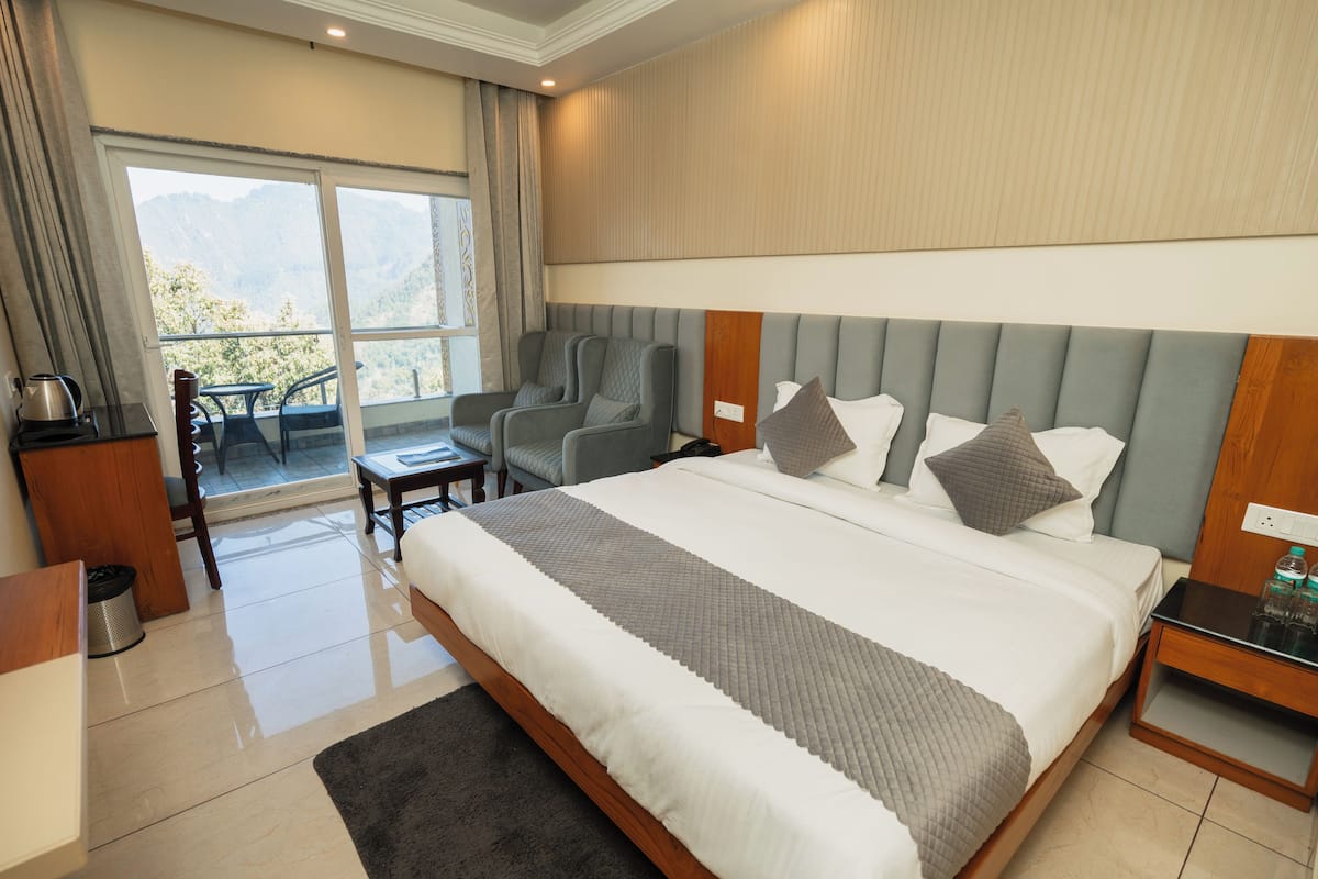 Habitación de hotel con una cama grande, una zona de estar con sillas, una mesita y vistas a las montañas a través de la ventana.