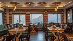 A restaurant with brick walls, large windows, and a view of a mountain. Tables and chairs are arranged for dining. - Trideus Chandseena Valley Resort (Dehradun)