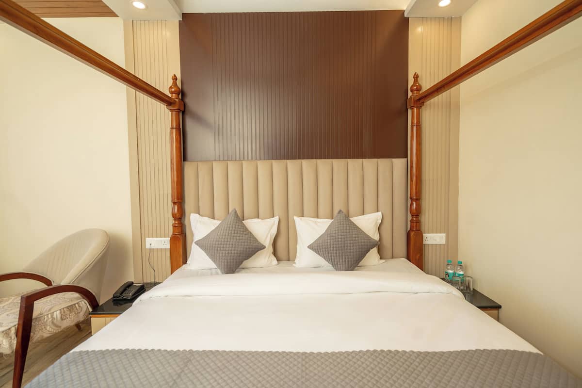 Habitación de hotel con una cama grande, cabecera de madera, mesita de noche con un teléfono y botellas de agua, y una silla.