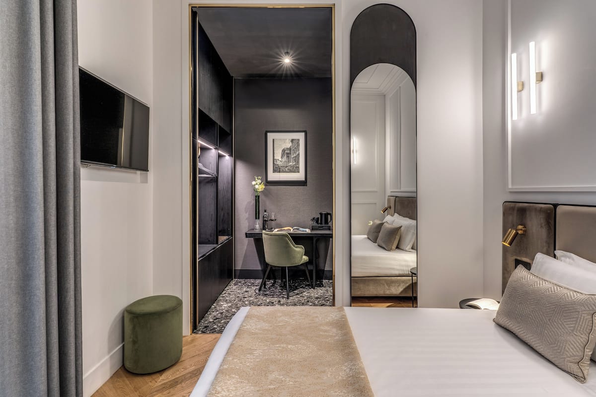 A modern hotel room with a large bed, a desk with a chair, a small green ottoman, and a framed picture on the wall.