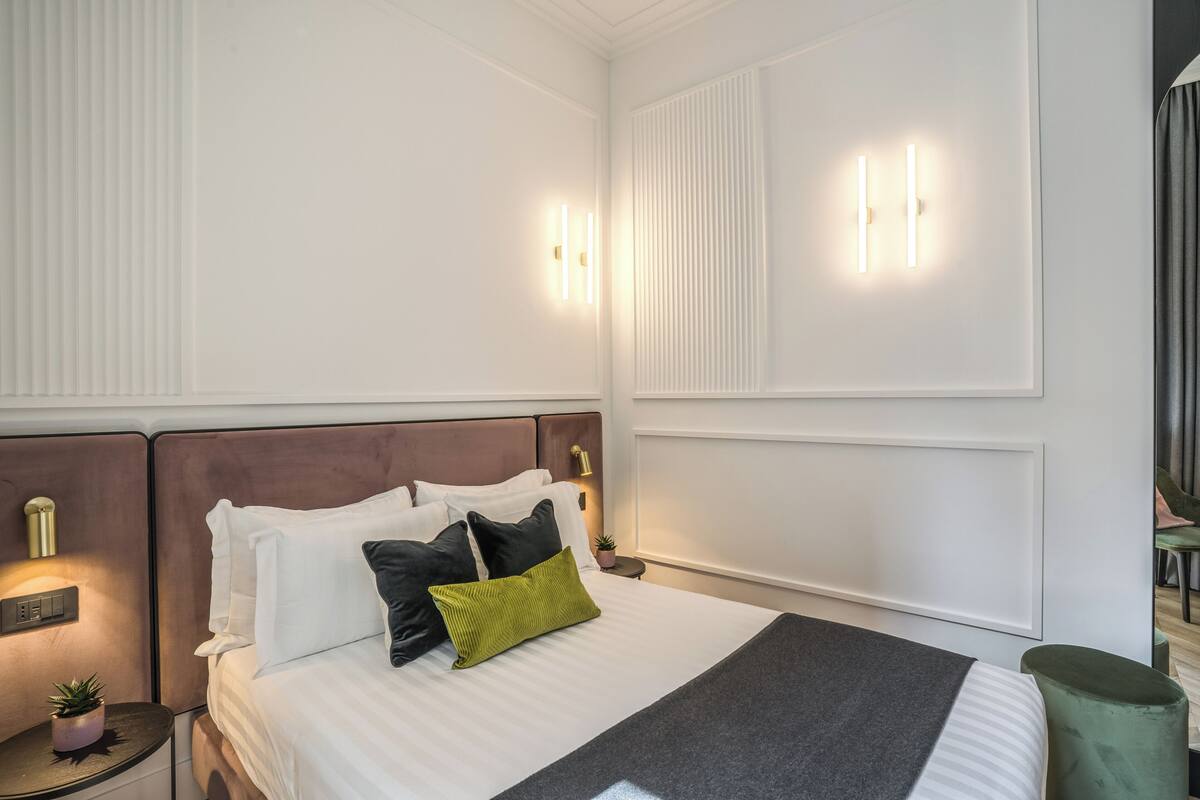 A neatly made bed with white and black pillows, a green accent pillow, and a grey throw blanket. There are two wall-mounted light fixtures on either side of the bed.