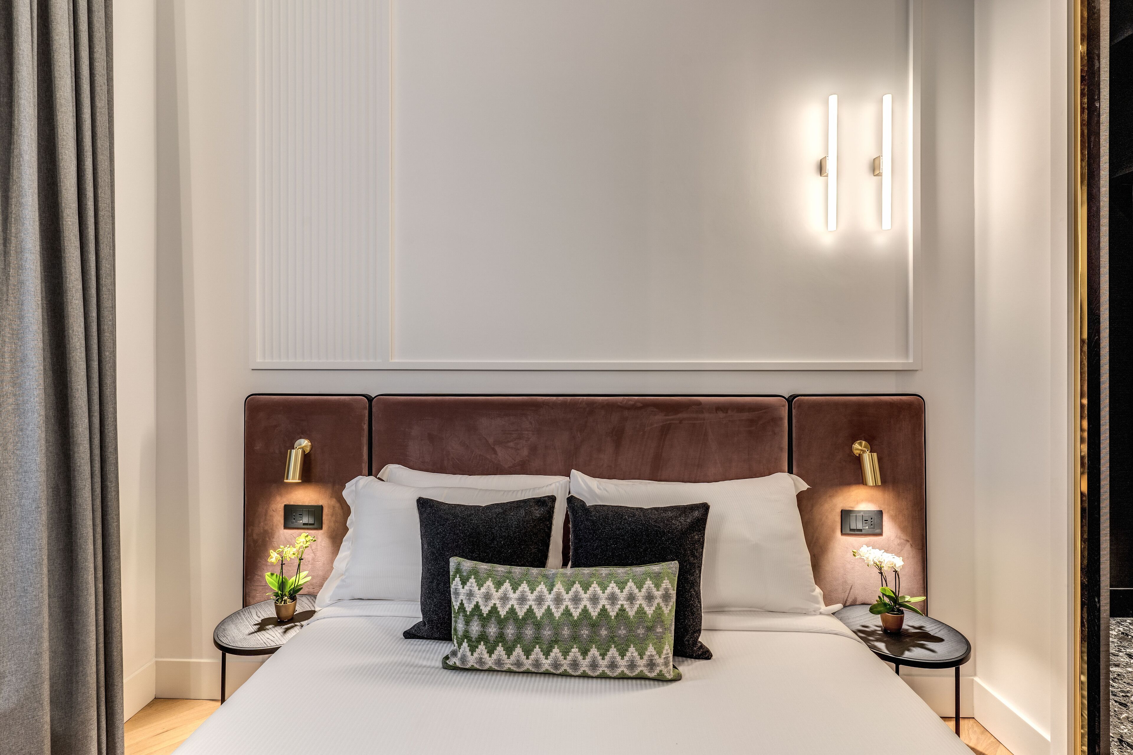A neatly made bed with a patterned pillow, two bedside tables with lamps, and a wall-mounted light fixture.