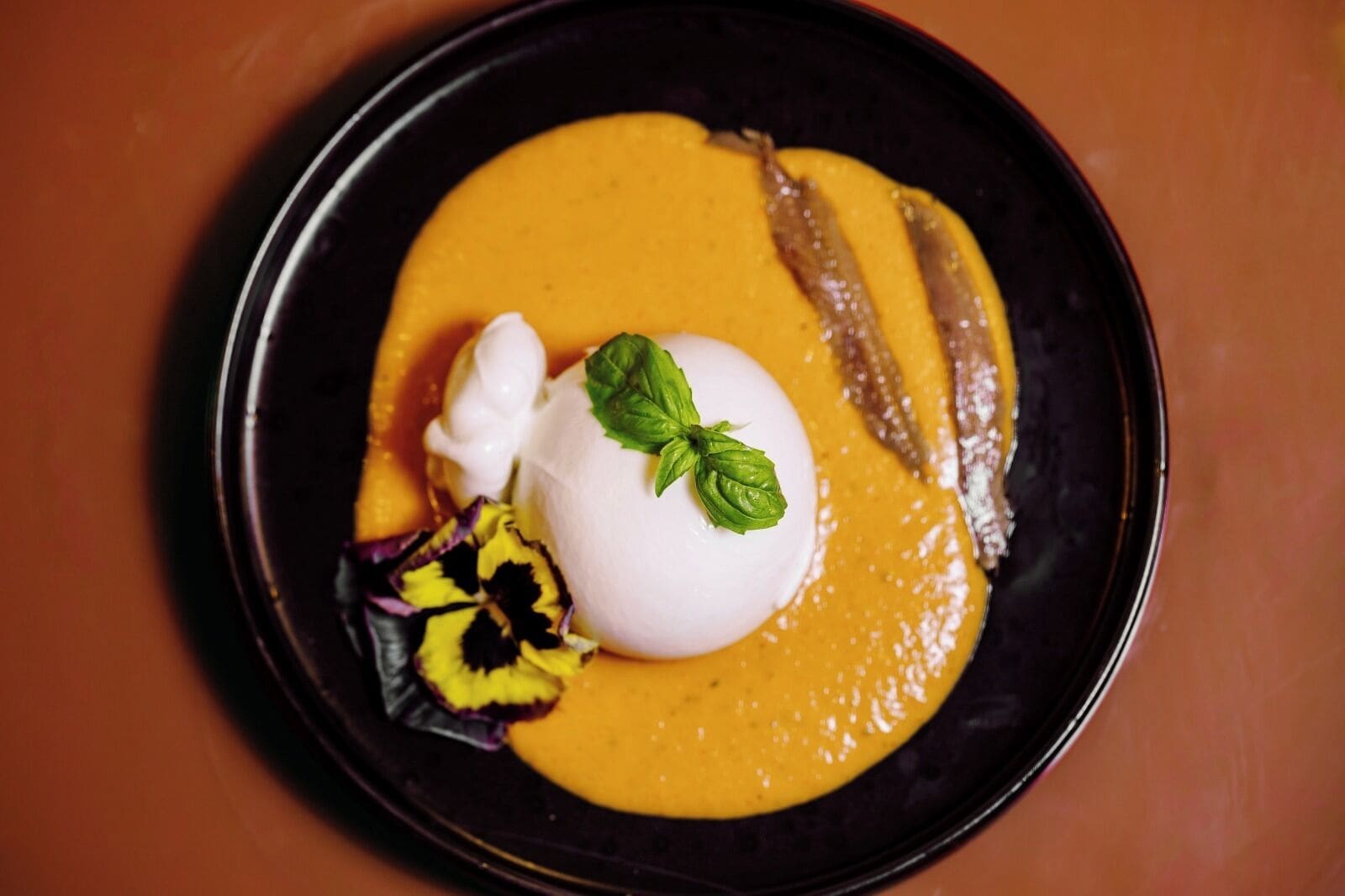 Un plato con sopa cremosa de naranja, una cucharada de crema, una hoja de albahaca y una rodaja de piña.