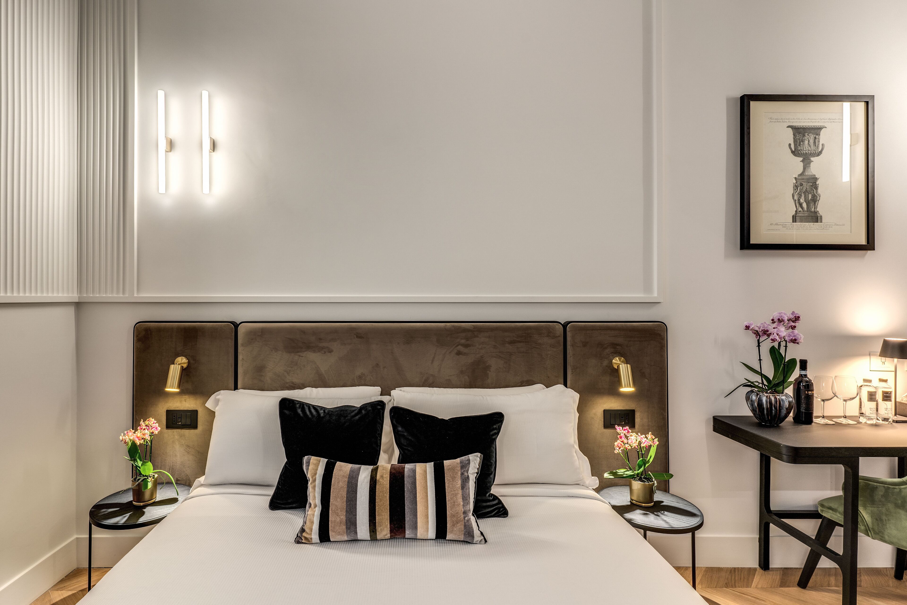 A neatly arranged bedroom with a large bed, bedside tables with lamps, a small table with a vase and flowers, and a framed artwork on the wall.