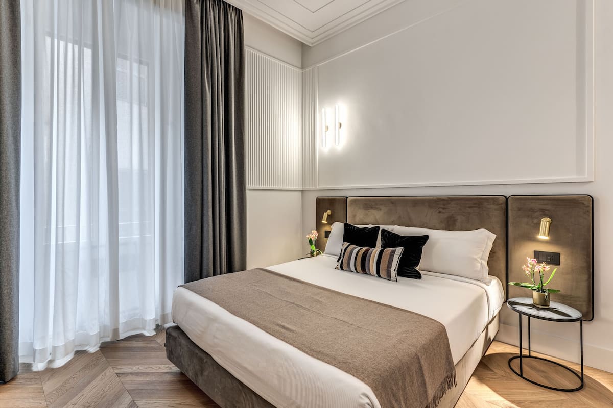 A neatly arranged hotel room with a large bed, a bedside table with a lamp, and a view of the outside through sheer curtains.