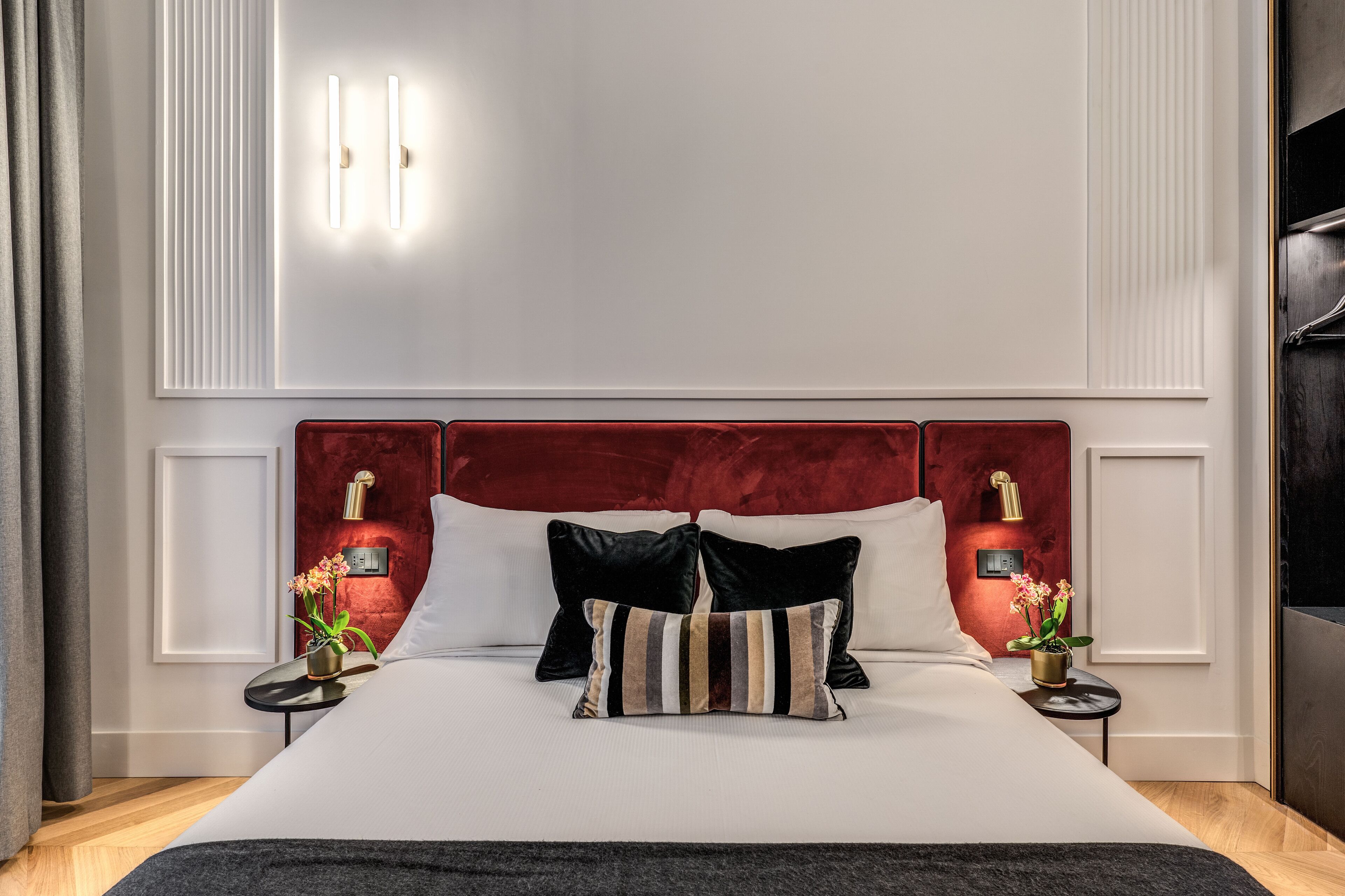 A neatly made bed with a red headboard, two bedside tables with lamps, and a potted plant on each side.