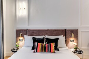 Una cama bien tendida con un cojín a rayas, dos mesitas de noche con lámparas y un aplique de pared.