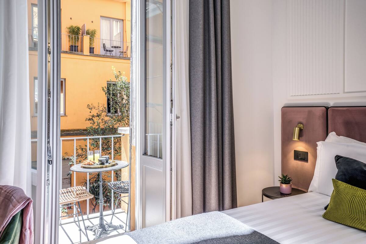 A hotel room with a balcony, a bed with pillows, a small table with a plant, and a window with curtains.