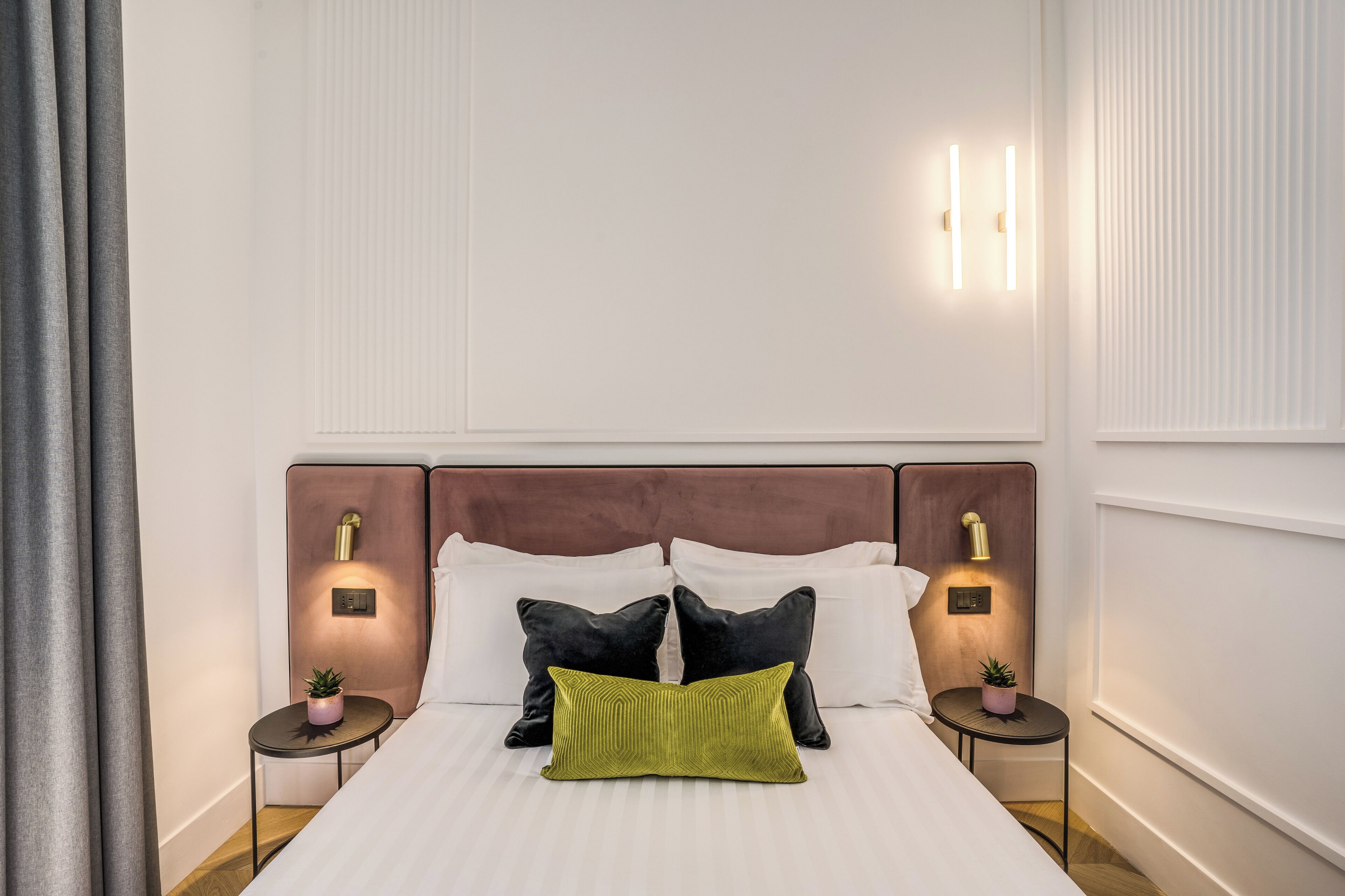Una cama bien hecha con un cabecero de madera, dos mesitas de noche con lámparas y una pequeña planta en maceta sobre cada una.