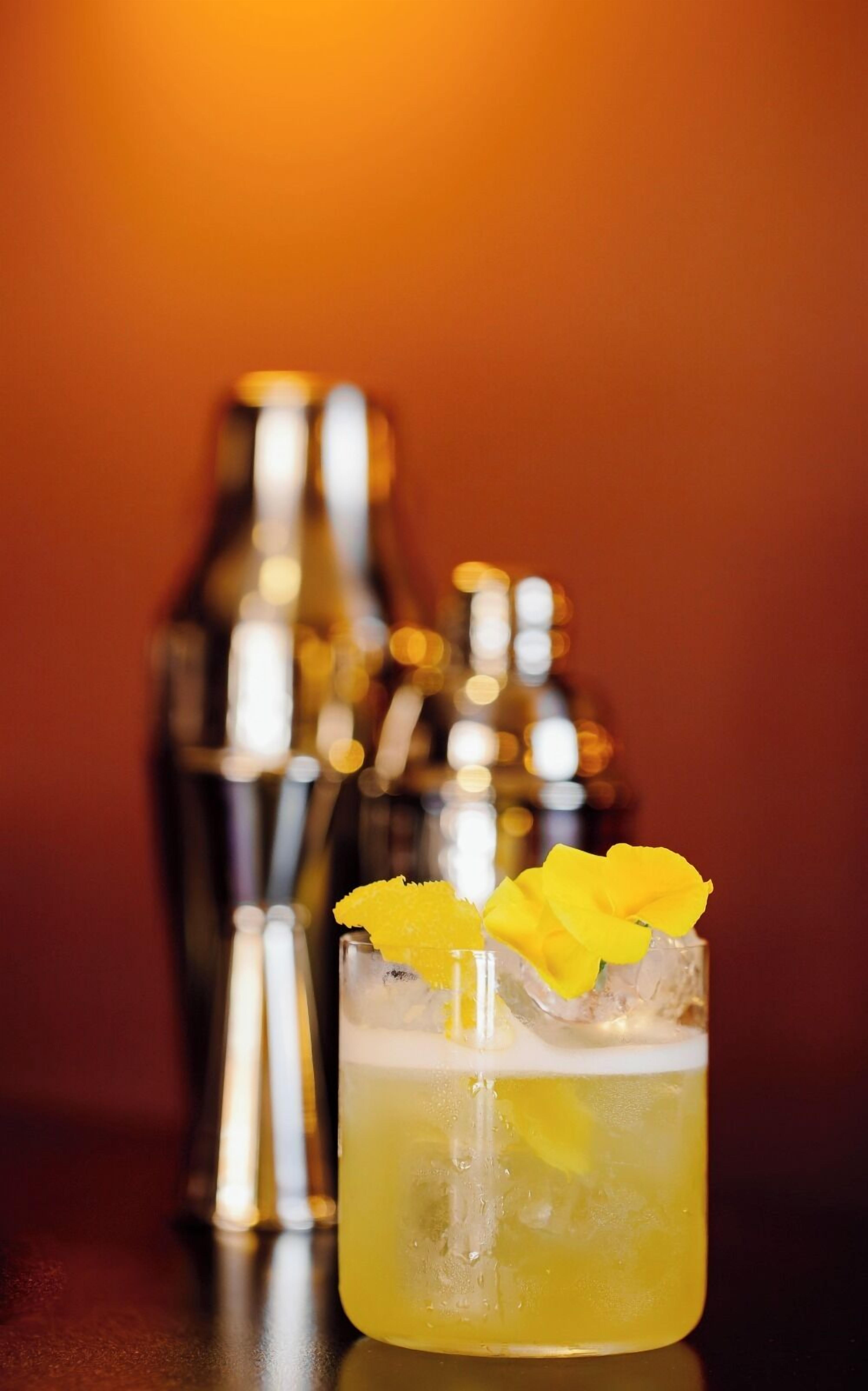 Een cocktailglas met een geel drankje, versierd met een gele bloem en sinaasappelschil, en ijsblokjes. Op de achtergrond staan twee cocktailshakers.