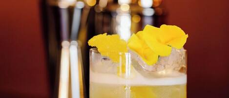 Een cocktailglas met een geel drankje, versierd met een gele bloem en sinaasappelschil, en ijsblokjes. Op de achtergrond staan twee cocktailshakers.