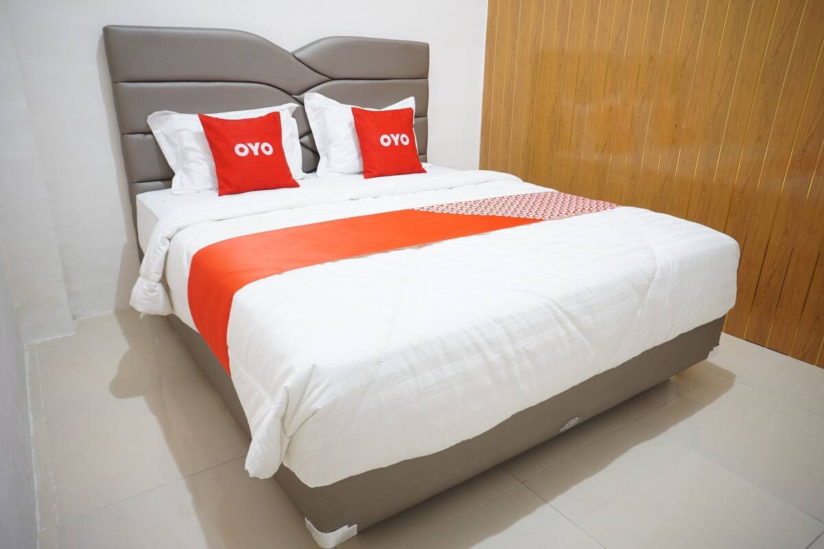 Una cama bien tendida con cabecera gris, ropa de cama blanca y roja, y dos almohadas con el texto "OYO".