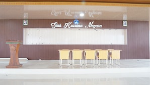 Een vergaderzaal met een spreekgestoelte, een lange tafel en een bordje met de tekst 'Smt. Kusuma Negara'.