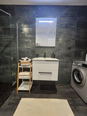 A modern bathroom with a glass shower enclosure, a built-in sink with a mirror, a white vanity, and a front-loading washing machine.