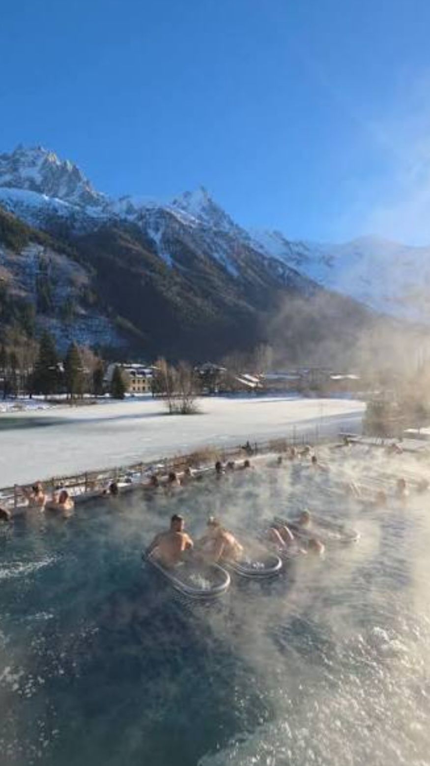 Une zone de sources chaudes où des personnes se détendent dans l’eau, entourées de montagnes enneigées et sous un ciel dégagé.
