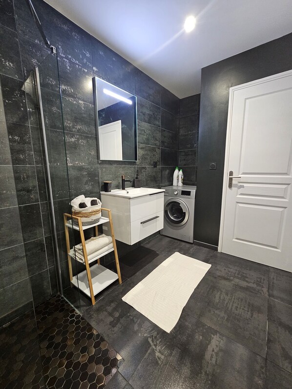 Une salle de bain moderne avec une douche à parois de verre, un lavabo blanc, un lave-linge et une étagère en bois où sont posées des serviettes.
