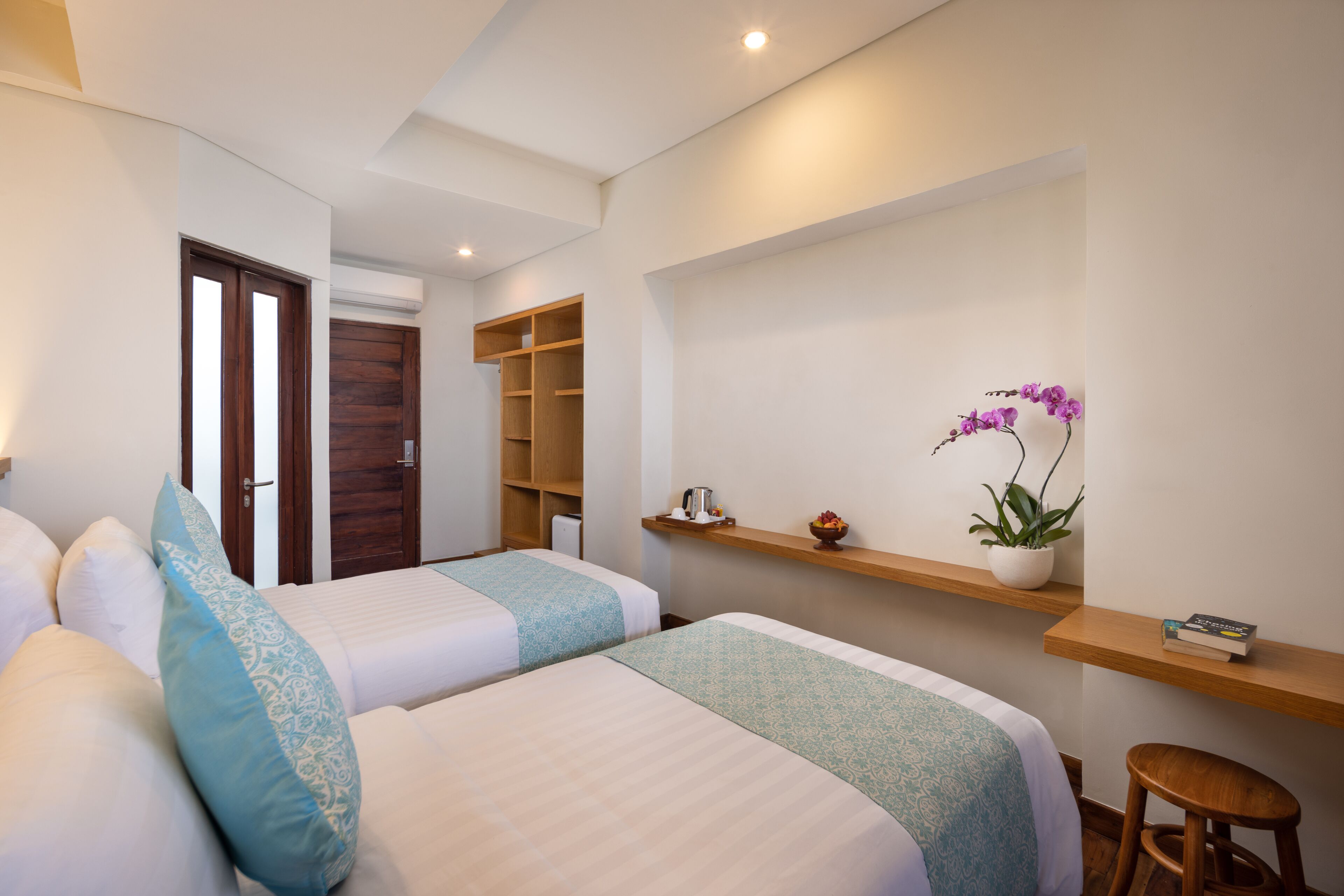 Een hotelkamer met twee bedden, een houten deur, een plankje met een plant erop en een klein krukje.