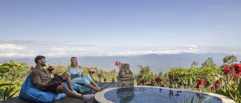 Two people sitting on a wooden deck by a hot tub with a stone statue and flowers, overlooking a mountainous landscape.