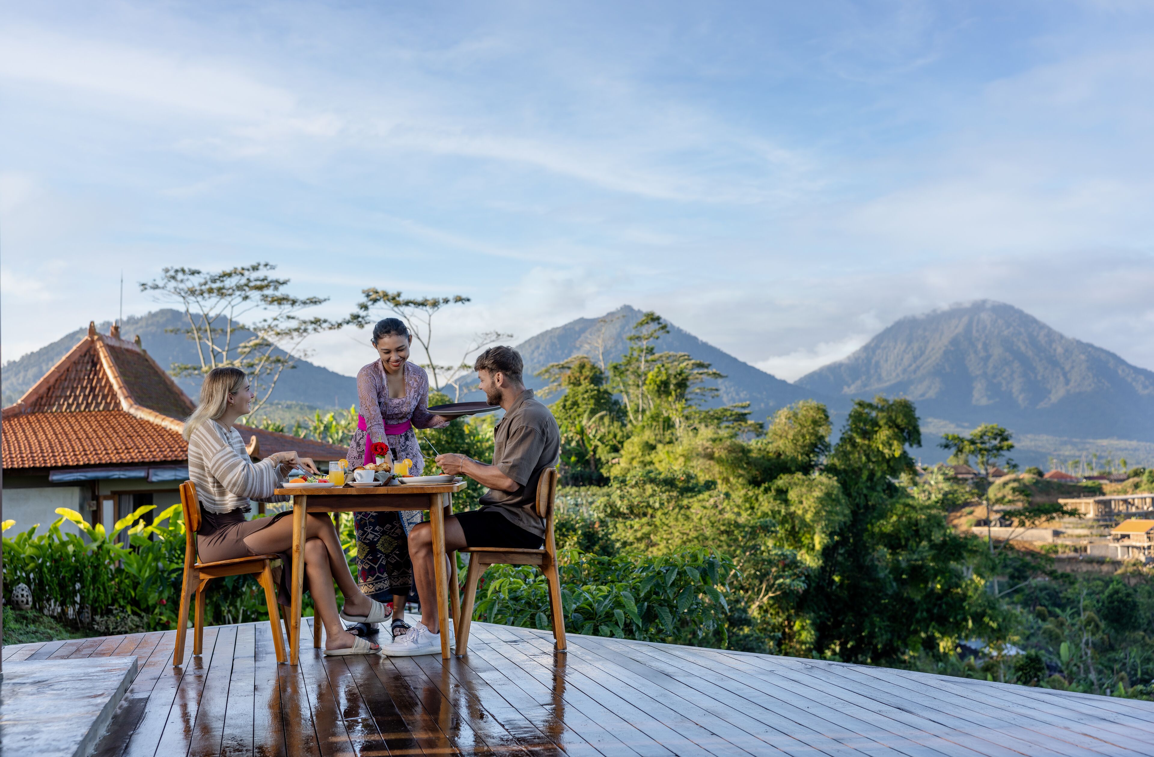 Una terraza de madera con una mesa donde tres personas están sentadas, disfrutando de una comida.