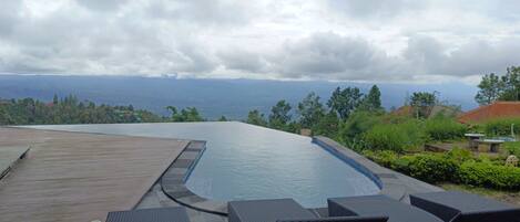 Una piscina infinita con terraza de madera y sillones de mimbre, con vistas a un paisaje montañoso.