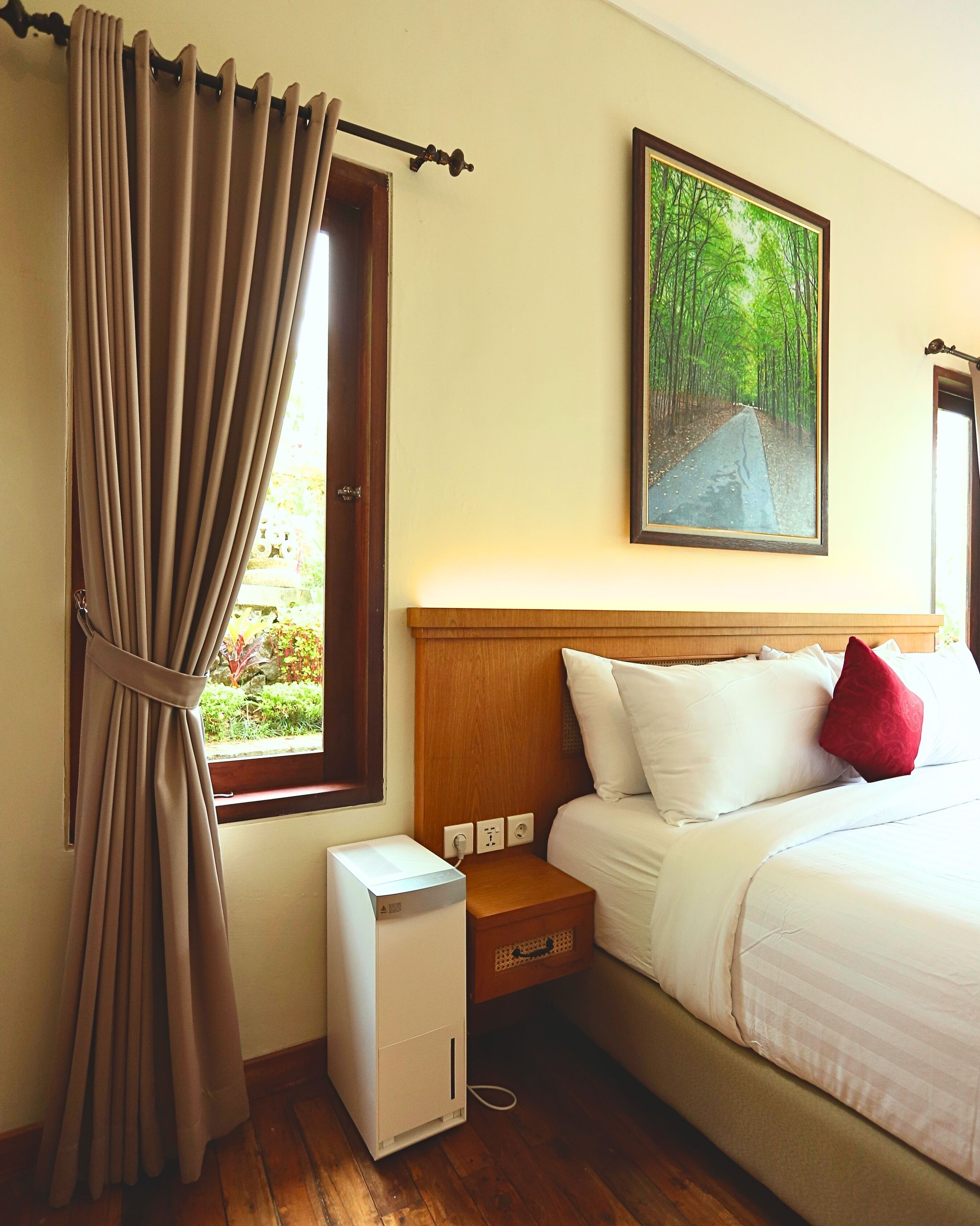 A hotel room with a bed, a nightstand, a small white heater, a window with curtains, and a painting of a forest path.