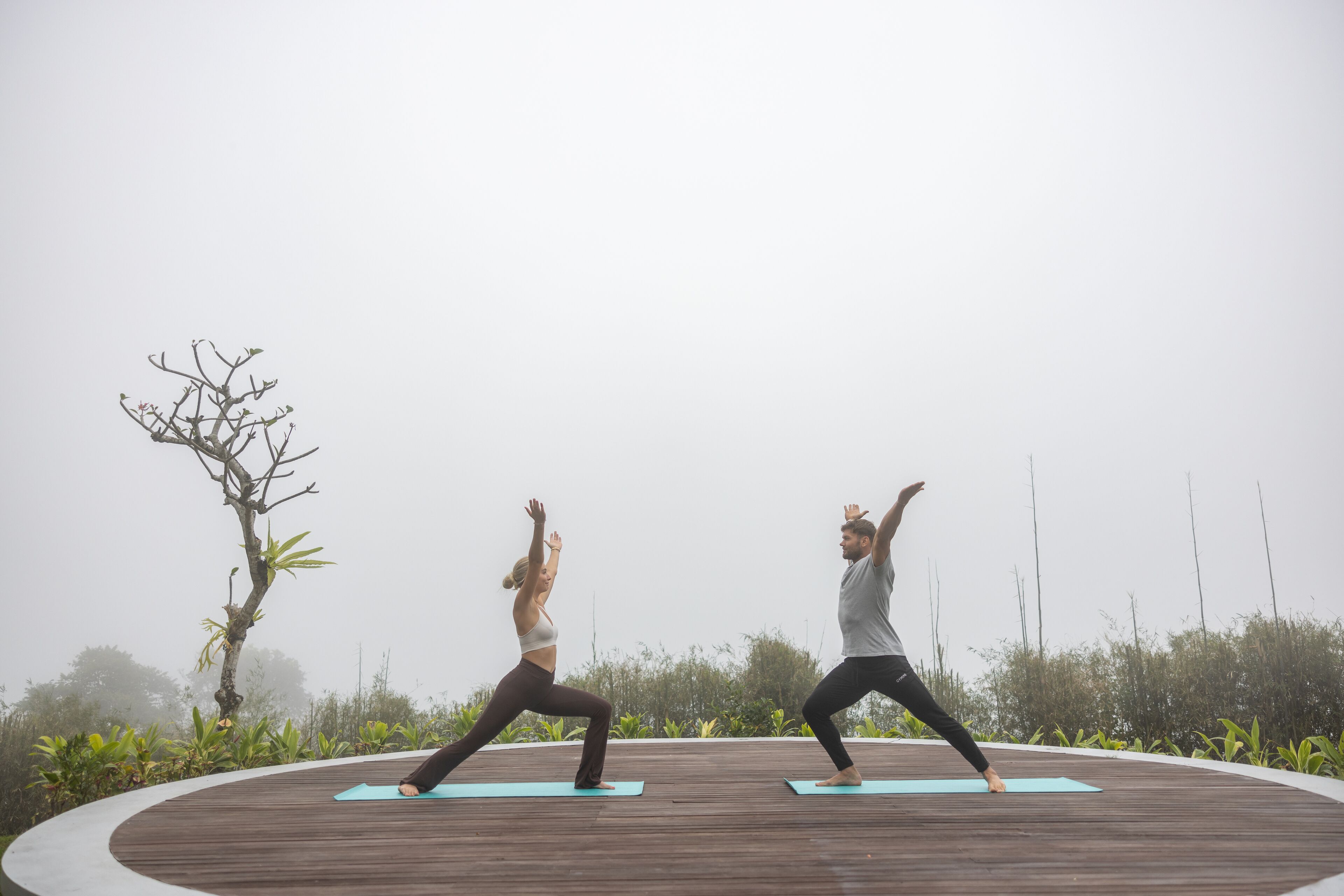 Two people performing yoga on a wooden deck with a misty background.