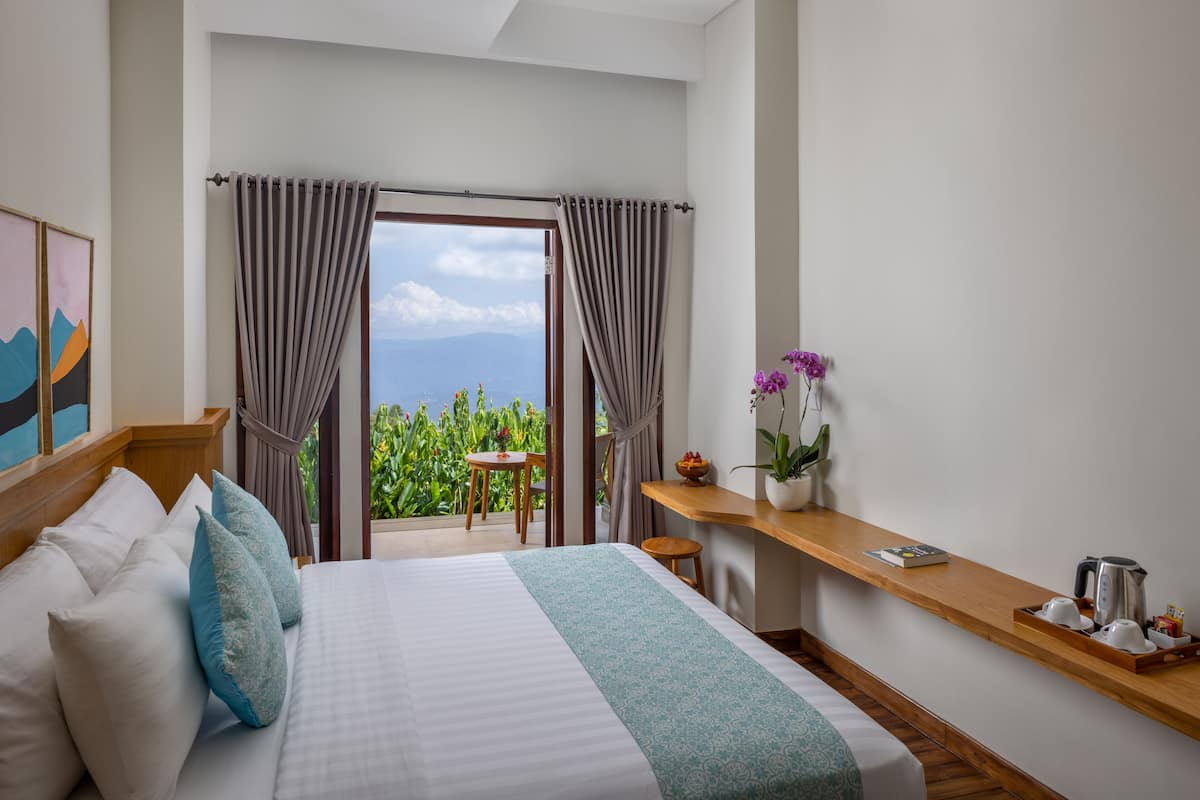 A hotel room with a large bed, a wooden nightstand, a small table, and a view of the outdoors.