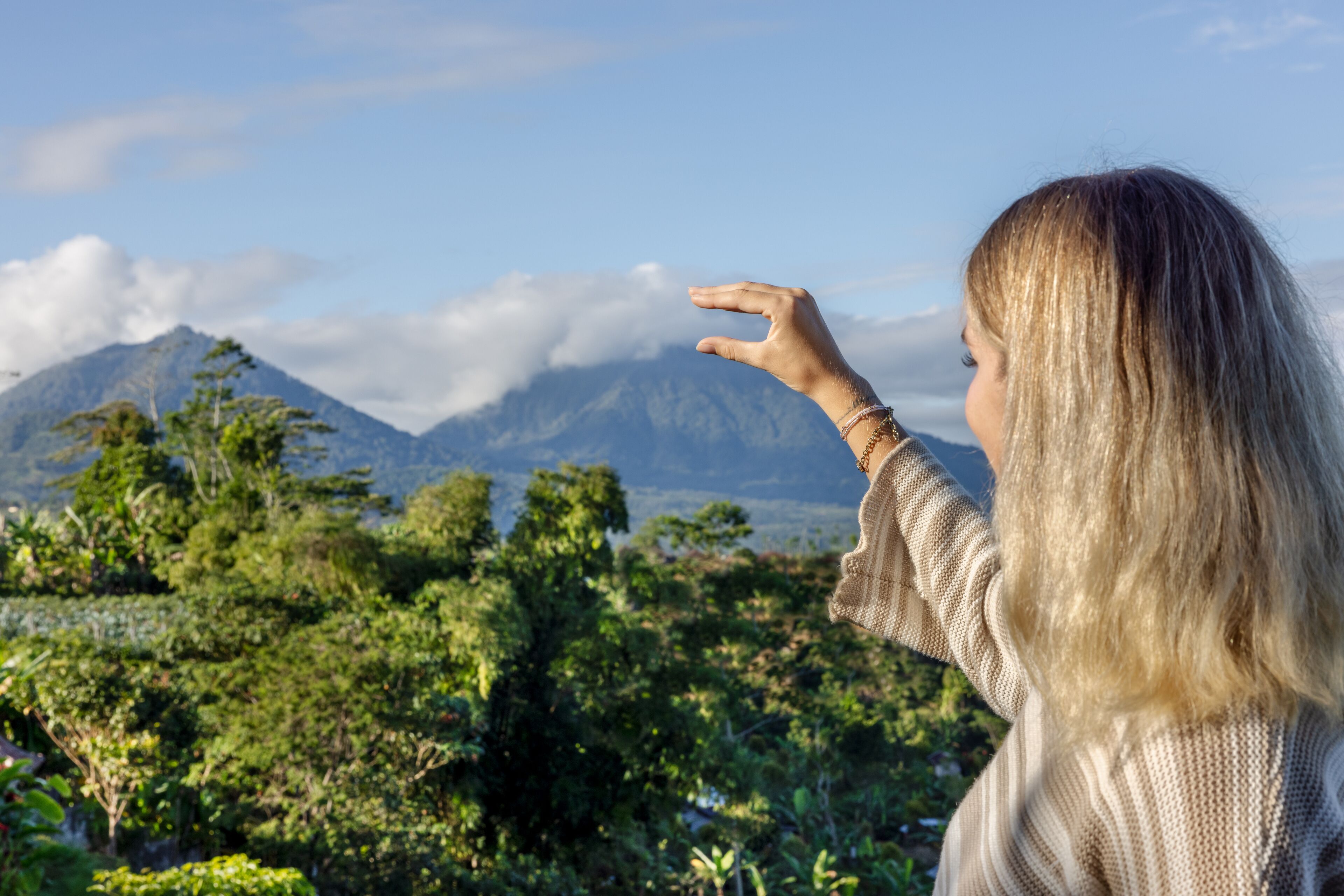 Une personne aux longs cheveux blonds, debout à l’extérieur, face à un paysage montagneux caractérisé par une végétation luxuriante et un ciel dégagé.