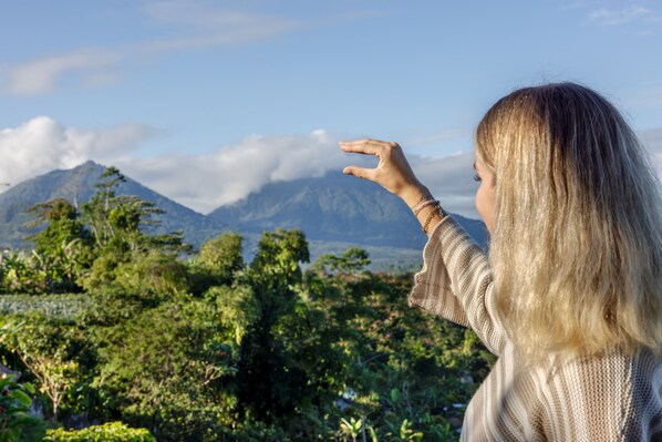 Una persona de cabello rubio largo parada al aire libre, frente a un paisaje montañoso con abundante vegetación y cielo despejado.