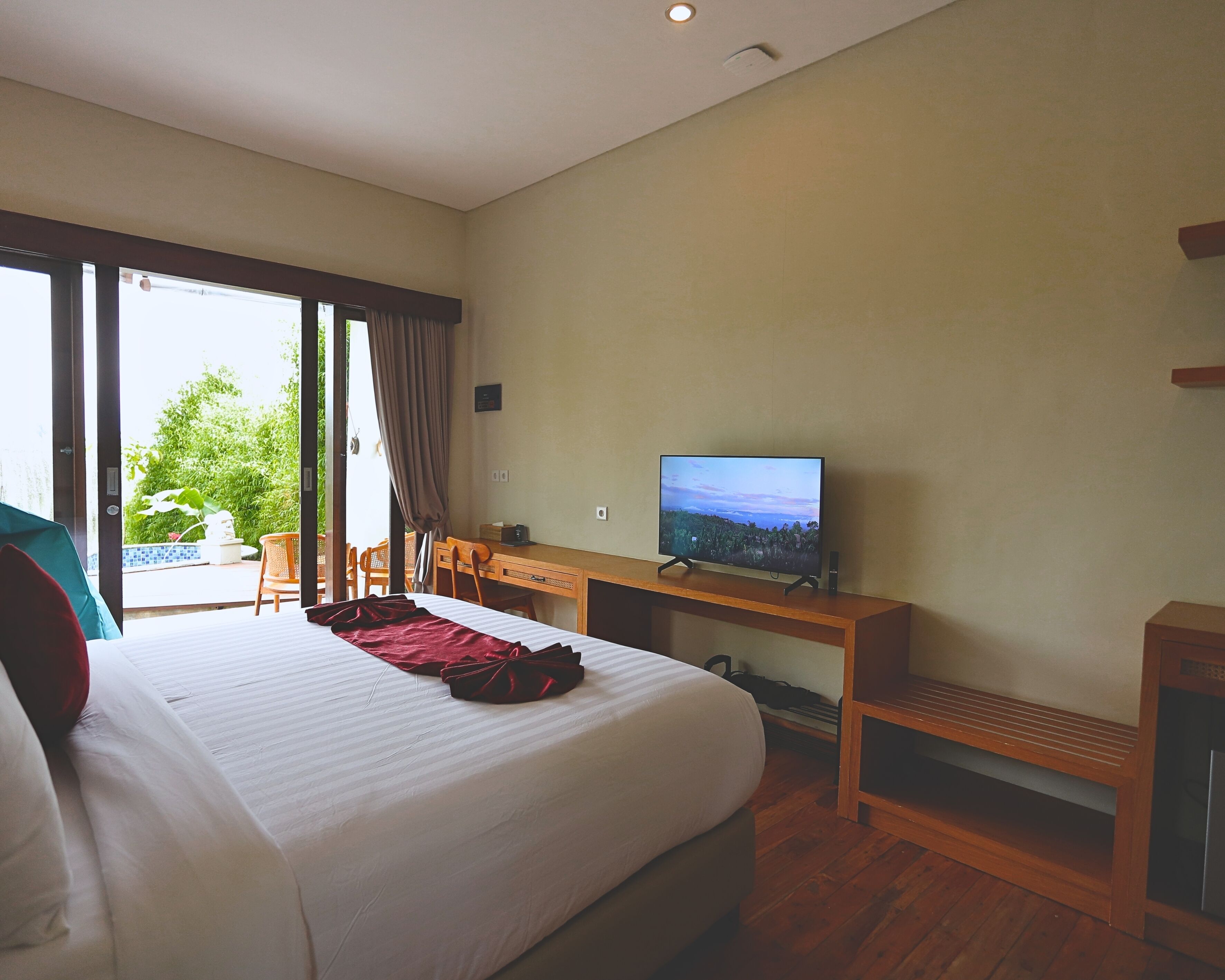 A hotel room with a large bed, a desk with a television, and a view of greenery outside.