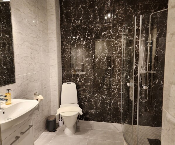 A modern bathroom with a toilet, sink, and glass-enclosed shower. - Brodderud Apartments (Mariestad)