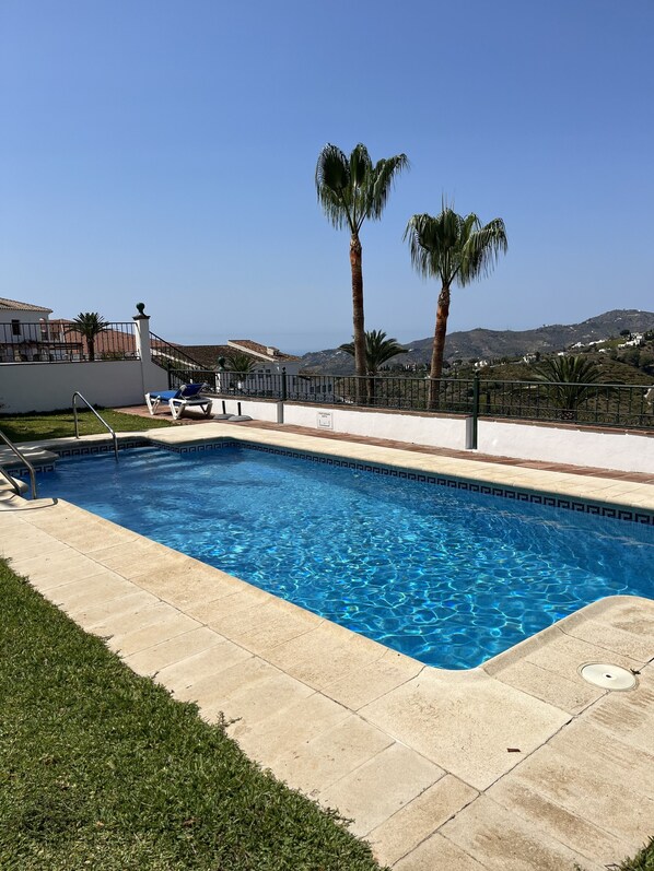 Una piscina rectangular con agua azul cristalina, rodeada por un solado de baldosas. Se ven dos palmeras y un edificio blanco al fondo.