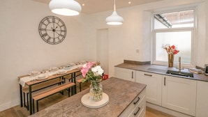 A modern kitchen with a dining area, featuring a clock, pendant lights, and a vase with flowers.
