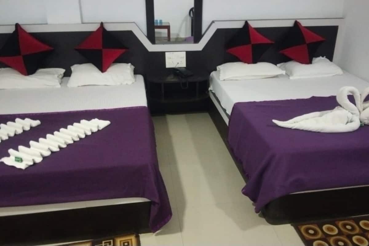 A hotel room with two beds, each with a purple bedspread and white pillows. There are decorative pillows with red and black patterns on the headboards. A mirror is mounted on the wall above the beds.