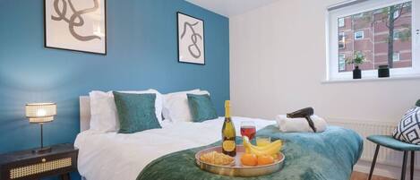 Un dormitorio con una cama, una mesita de noche con lámpara, una bandeja con frutas y una botella, y dos cuadros en la pared.