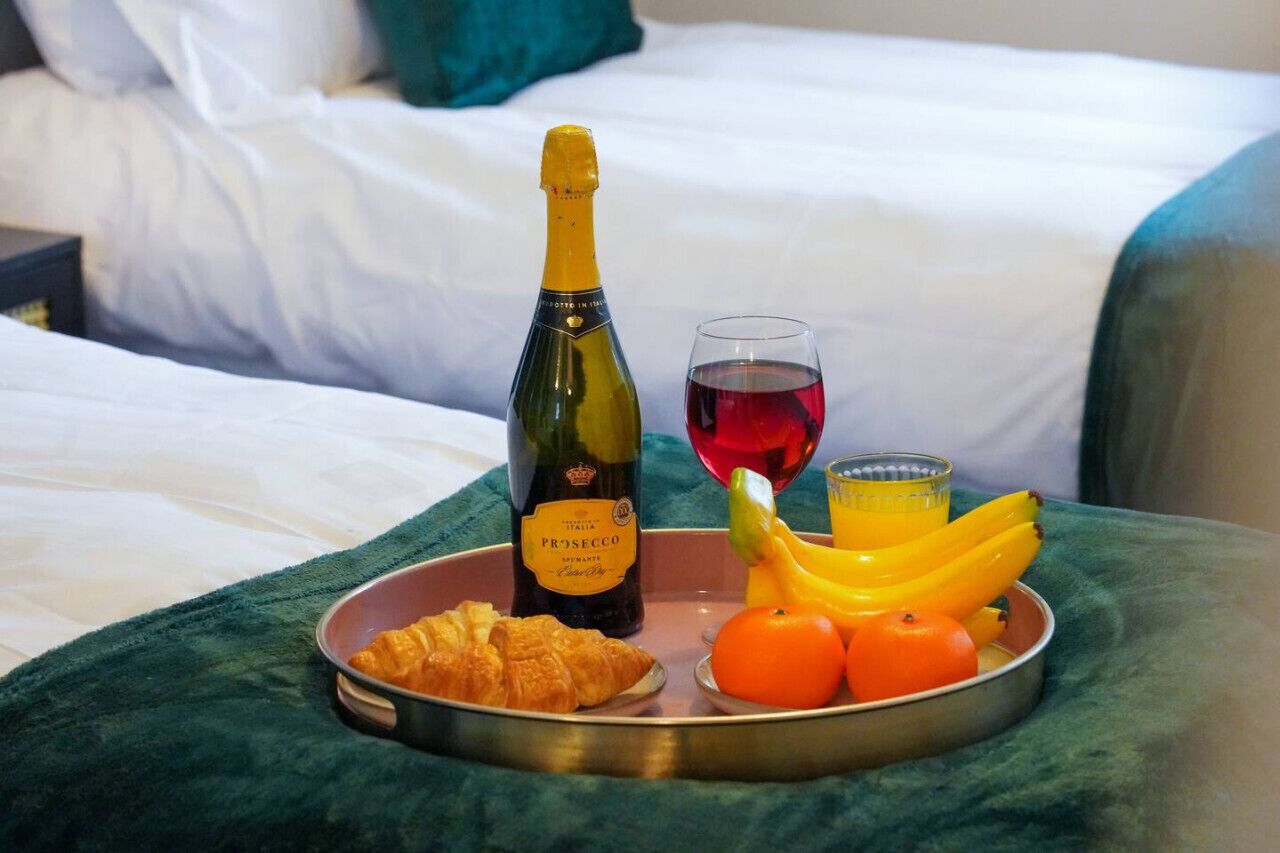 A tray with a bottle of Prosecco, a glass of red wine, and a selection of fruits and croissants on a bed.