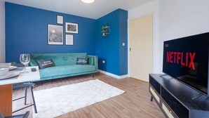 Ein Wohnzimmer mit einem türkisfarbenen Sofa, einem weißen Teppich, einem Fernseher mit der Aufschrift „NETFLIX“ und einer Wand mit gerahmten Bildern.
